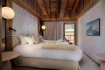 Experience luxury mountain living in this beautifully appointed bedroom, featuring a plush king-sized bed, warm wood accents, and a stunning view of snow-capped peaks through the balcony door.