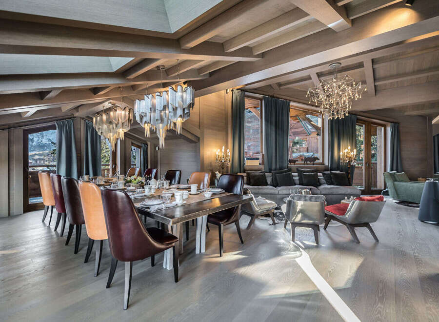 Experience luxurious mountain living in this stunning dining and living area, featuring a grand wooden dining table set for a feast and elegant chandeliers that illuminate the space.
