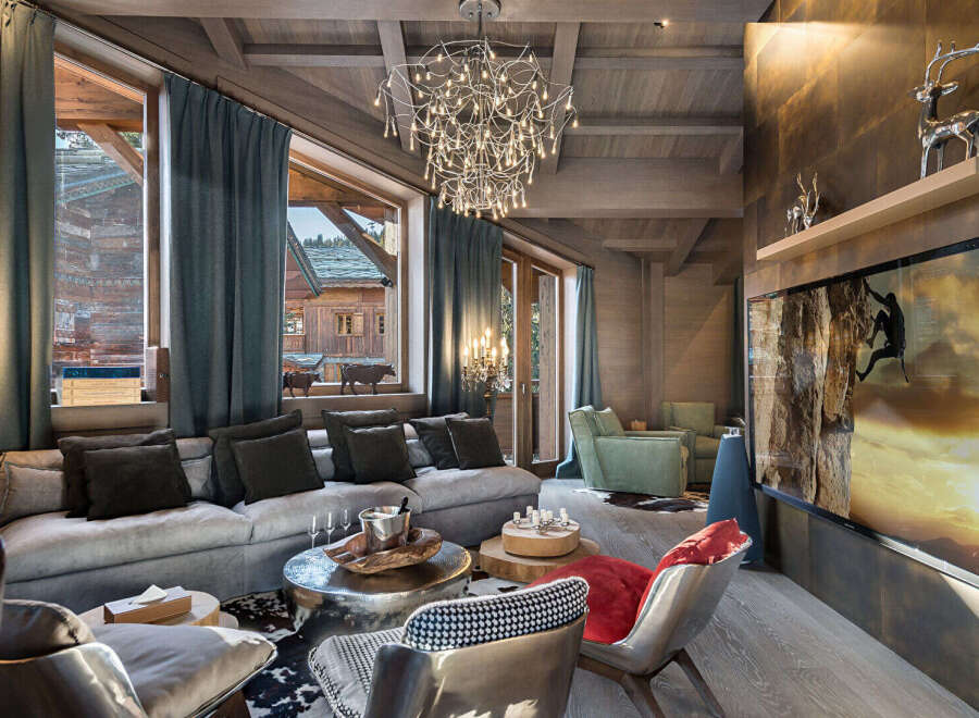 Experience luxurious mountain living in this stunning chalet living room, featuring a plush sectional sofa, a striking modern chandelier, and breathtaking views of a charming village through expansive windows.