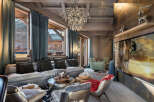 Experience luxurious mountain living in this stunning chalet living room, featuring a plush sectional sofa, a striking modern chandelier, and breathtaking views of a charming village through expansive windows.