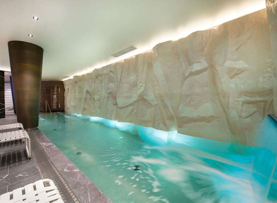 Experience ultimate relaxation in this luxurious indoor pool area, featuring a stunning rock-like wall with integrated waterfall and soothing blue lighting.