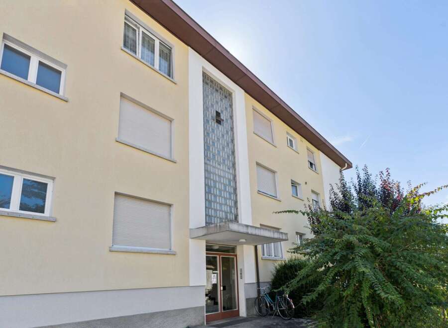 This apartment building features a light yellow facade with multiple windows, some equipped with roller shutters for privacy and light control.