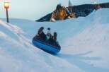 Experience winter wonderland fun with this thrilling snow tubing adventure! Glide down snowy slopes on a blue inflatable tube, with a whimsical giant troll statue and a castle-like structure in the background adding to the magical atmosphere.