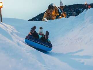Experience winter wonderland fun with this thrilling snow tubing adventure! Glide down snowy slopes on a blue inflatable tube, with a whimsical giant troll statue and a castle-like structure in the background adding to the magical atmosphere.