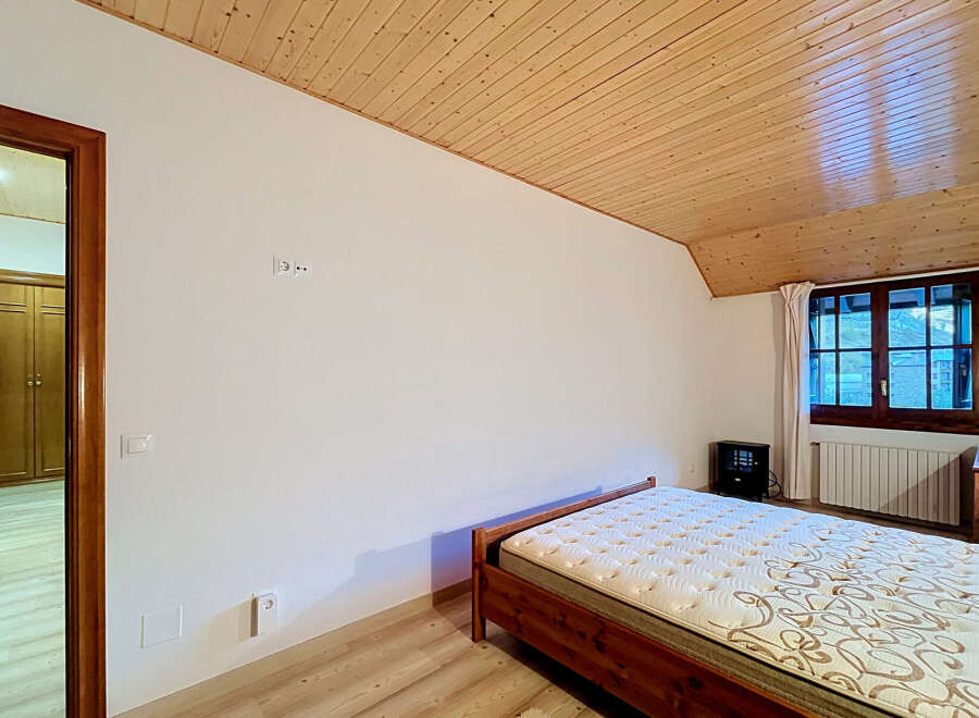 This charming bedroom features a comfortable wooden bed with a patterned mattress, set against a backdrop of white walls and a warm, slatted wooden ceiling.