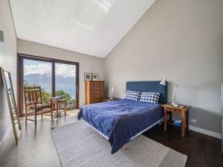Experience breathtaking mountain and lake views from this serene bedroom, featuring a comfortable blue bed with gingham pillows and a spacious wooden dresser.