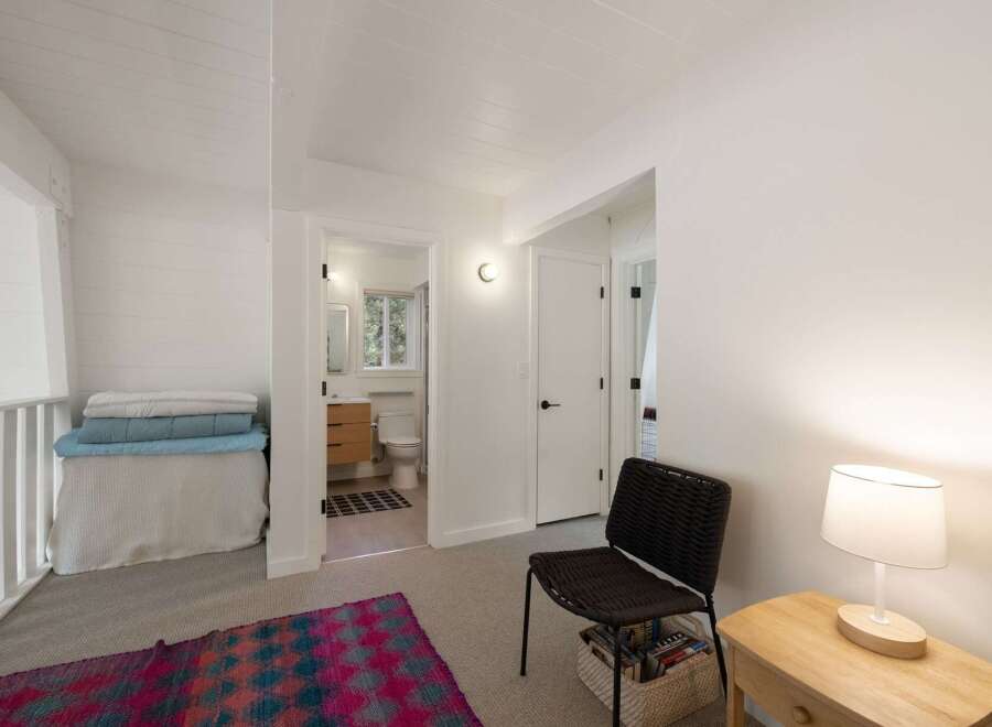 This bright and airy bedroom features a cozy seating area with a woven chair and a bedside table with a lamp.