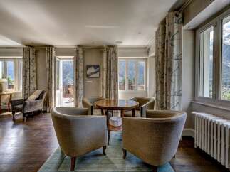 Experience breathtaking mountain views from this elegant living space, featuring a cozy seating area with plush armchairs and a central wooden table.