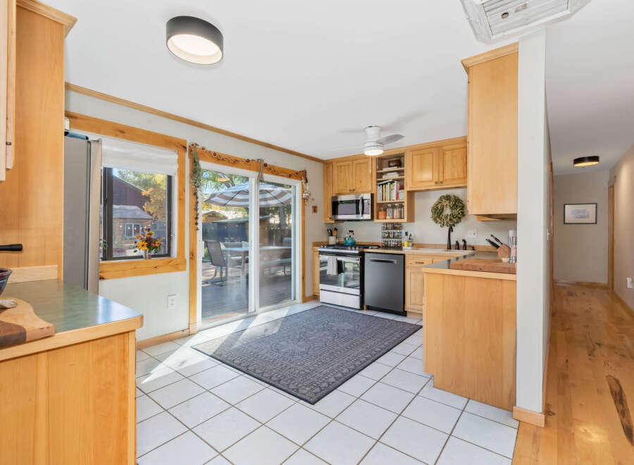 This bright and airy kitchen features light wood cabinetry, white tiled floors, and a sliding glass door that opens to a deck and outdoor seating area.
