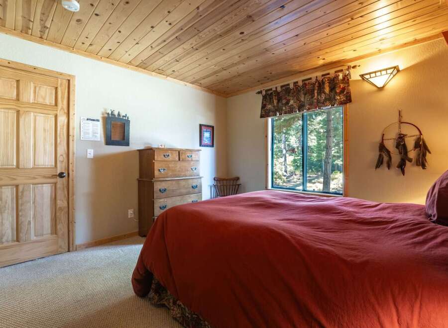 Experience rustic charm in this cozy bedroom, featuring a warm wooden ceiling and a large window offering serene forest views.