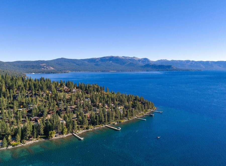 Experience the ultimate lakeside living with this stunning aerial view of homes nestled among pine trees along the crystal-clear waters of Lake Tahoe.