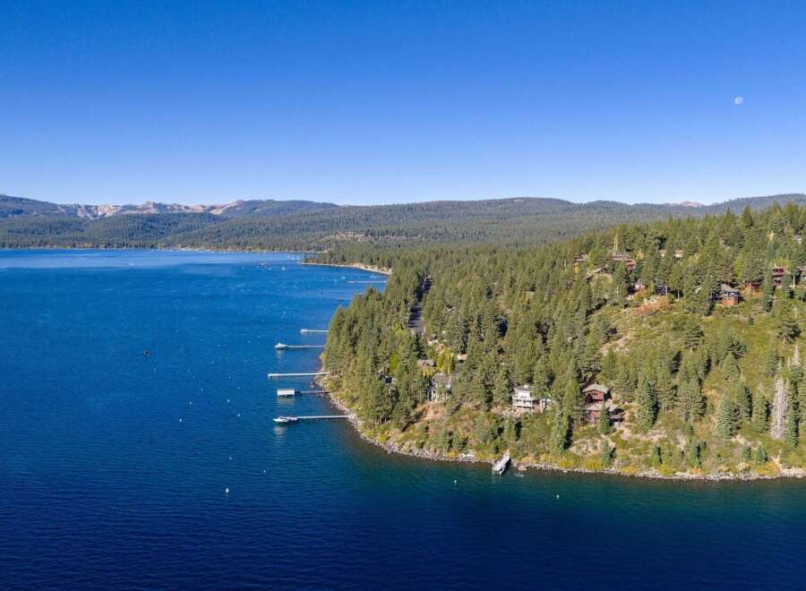 Experience luxury living with this stunning aerial view of waterfront properties nestled among lush pine trees on a hillside overlooking a vast, deep blue lake.