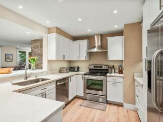 This modern kitchen boasts sleek white cabinetry and stainless steel appliances, including a refrigerator, dishwasher, and oven with a matching range hood.