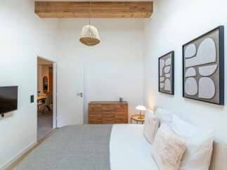 This modern bedroom features a comfortable bed with plush pillows and a soft grey throw, complemented by abstract wall art and a rustic wooden dresser.