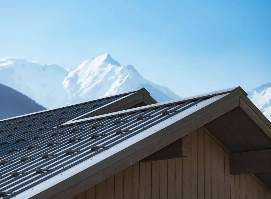 Experience luxury mountain living with this stunning property featuring a modern metal roof and rustic wooden siding, perfectly complementing the majestic snow-capped peaks in the background.
