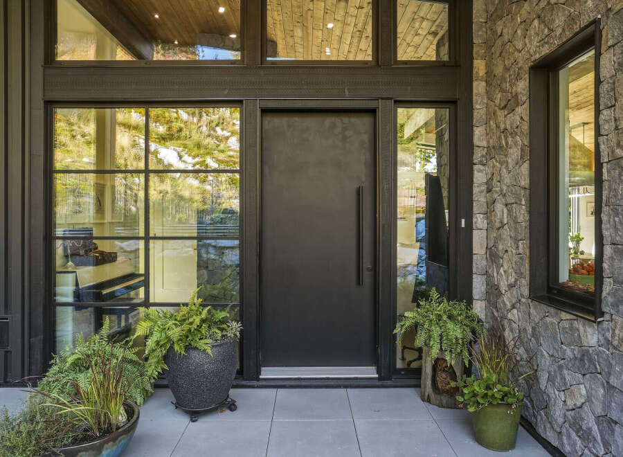 This modern home features a striking entrance with a large, dark metal door flanked by expansive windows that offer views of the surrounding natural landscape.