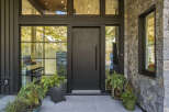 This modern home features a striking entrance with a large, dark metal door flanked by expansive windows that offer views of the surrounding natural landscape.