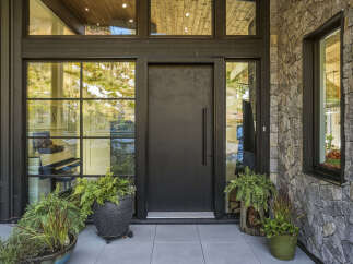This modern home features a striking entrance with a large, dark metal door flanked by expansive windows that offer views of the surrounding natural landscape.