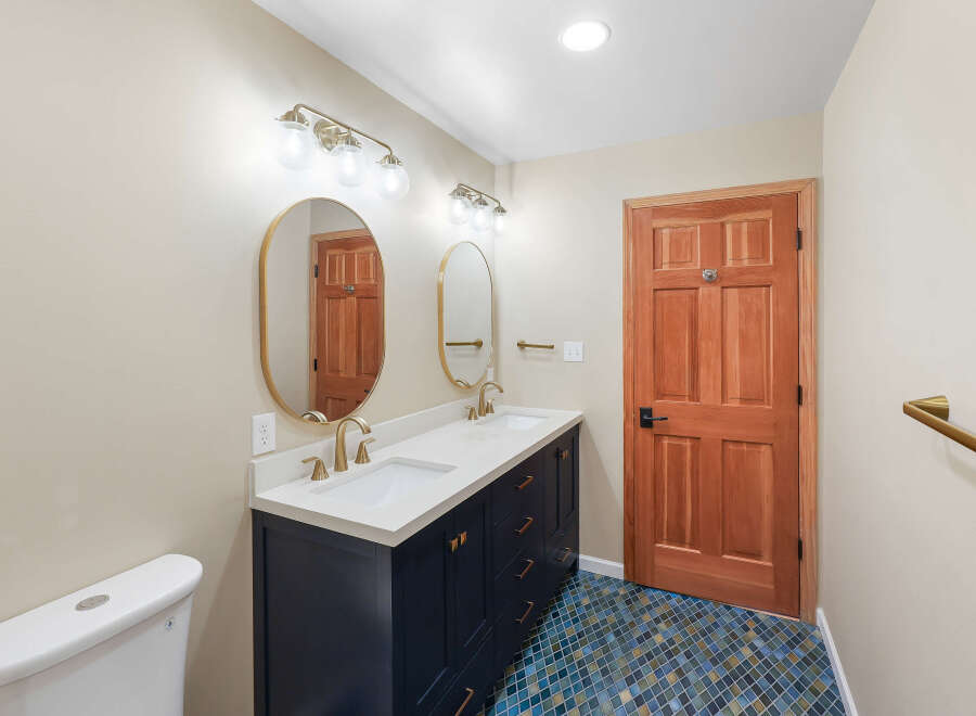 This bathroom features a spacious double vanity with a white quartz countertop and a navy blue cabinet base, complemented by brushed gold fixtures and oval mirrors.