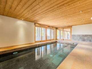 Experience luxury and relaxation in this indoor swimming pool area, featuring a sleek, dark-tiled pool that reflects the natural light streaming through large wooden-framed glass doors.