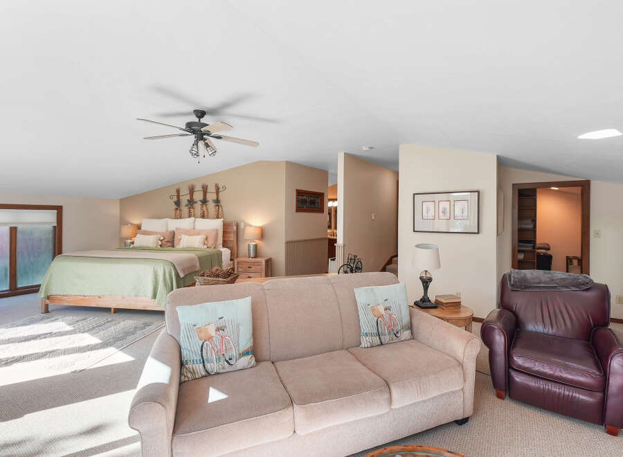This spacious loft bedroom offers a comfortable king-size bed with a light green quilt and decorative pillows, complemented by a cozy seating area featuring a beige sofa and a rich brown leather armchair.