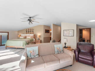 This spacious loft bedroom offers a comfortable king-size bed with a light green quilt and decorative pillows, complemented by a cozy seating area featuring a beige sofa and a rich brown leather armchair.