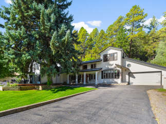Nestled amidst mature trees, this stunning white home offers a picturesque setting with a lush green lawn and a charming raised garden bed.