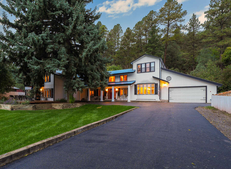 Nestled amidst towering pine trees, this stunning modern farmhouse boasts a crisp white exterior with black trim and a striking blue metal roof.