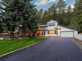 Nestled amidst towering pine trees, this stunning modern farmhouse boasts a crisp white exterior with black trim and a striking blue metal roof.