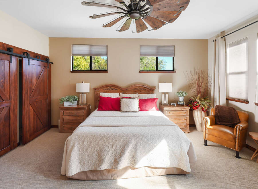 Experience rustic charm in this inviting bedroom featuring a grand wooden barn door and a striking windmill-inspired ceiling fan.