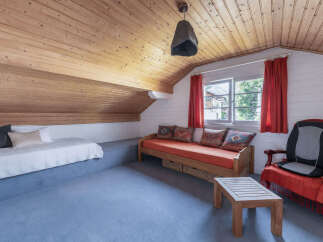 This charming attic room features a cozy sleeping area with a white quilted bed and decorative pillows, complemented by a rustic wooden daybed with vibrant orange cushions.