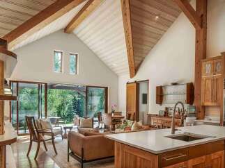 Experience rustic elegance in this spacious living area, featuring a vaulted ceiling with exposed wooden beams and a wall of glass doors that open to a lush, green outdoor space.