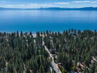 Experience the ultimate lakeside lifestyle with this stunning aerial view of a property nestled amongst towering pine trees, offering direct access to a pristine beach and the crystal-clear waters of Lake Tahoe.