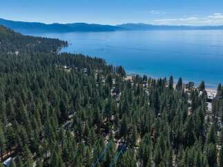 Experience the ultimate lakeside lifestyle with this stunning aerial view of a property nestled amidst a dense pine forest, offering direct access to a pristine beach and the sparkling blue waters of Lake Tahoe.