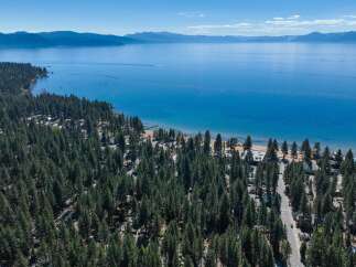 Experience the ultimate lakeside lifestyle with this stunning aerial view of a property nestled amongst towering pine trees, offering direct access to a pristine sandy beach and the crystal-clear waters of Lake Tahoe.