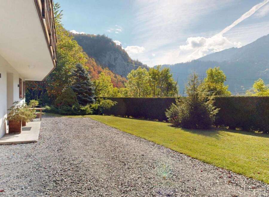 Experience the charm of this mountain retreat, featuring a white exterior with rustic wooden shutters and a balcony overlooking a lush green lawn and a gravel driveway.