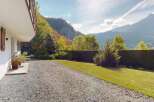 Experience the charm of this mountain retreat, featuring a white exterior with rustic wooden shutters and a balcony overlooking a lush green lawn and a gravel driveway.