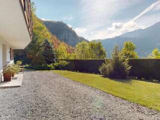 Experience the charm of this mountain retreat, featuring a white exterior with rustic wooden shutters and a balcony overlooking a lush green lawn and a gravel driveway.