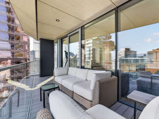 Experience luxurious urban living on this stunning balcony, featuring a spacious white sectional sofa and a chic wicker accent chair, perfect for enjoying breathtaking city views.
