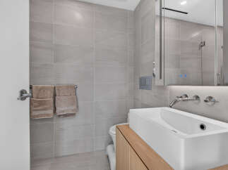 This modern bathroom features sleek grey tiled walls and floors, complemented by a minimalist white vessel sink atop a warm wood vanity.