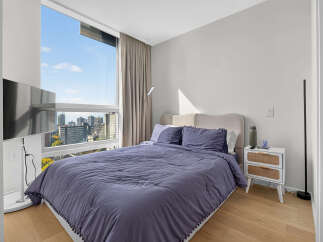 Experience urban living in this stylish bedroom featuring a plush bed with a textured headboard and a large window offering panoramic city views.