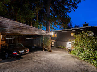 This property features a charming carport with a dark-colored Jaguar parked inside, nestled amongst lush greenery and tall trees.