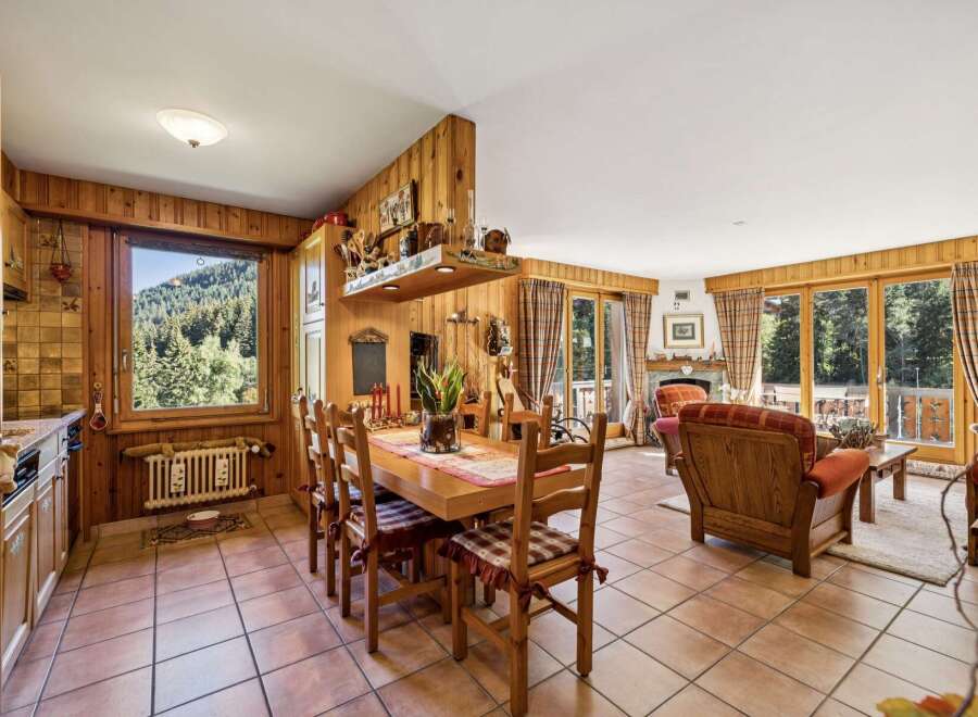 Experience cozy living in this charming mountain retreat, featuring a warm wood-paneled dining area with a large window offering stunning forest views.