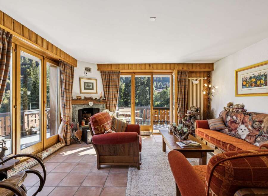 Experience cozy living in this charming mountain retreat, featuring a rustic stone fireplace and large wooden-framed windows that offer breathtaking views of the surrounding forest and valley.