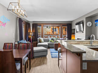 This open-concept living space features a modern dining area with a rich wood table and upholstered chairs, flowing seamlessly into a comfortable living room with a sectional sofa and vibrant accent pillows.