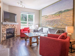 Cozy living room featuring a stone fireplace with a mounted TV, comfortable seating including a grey sofa and red accent chairs, and a rustic wooden coffee table.