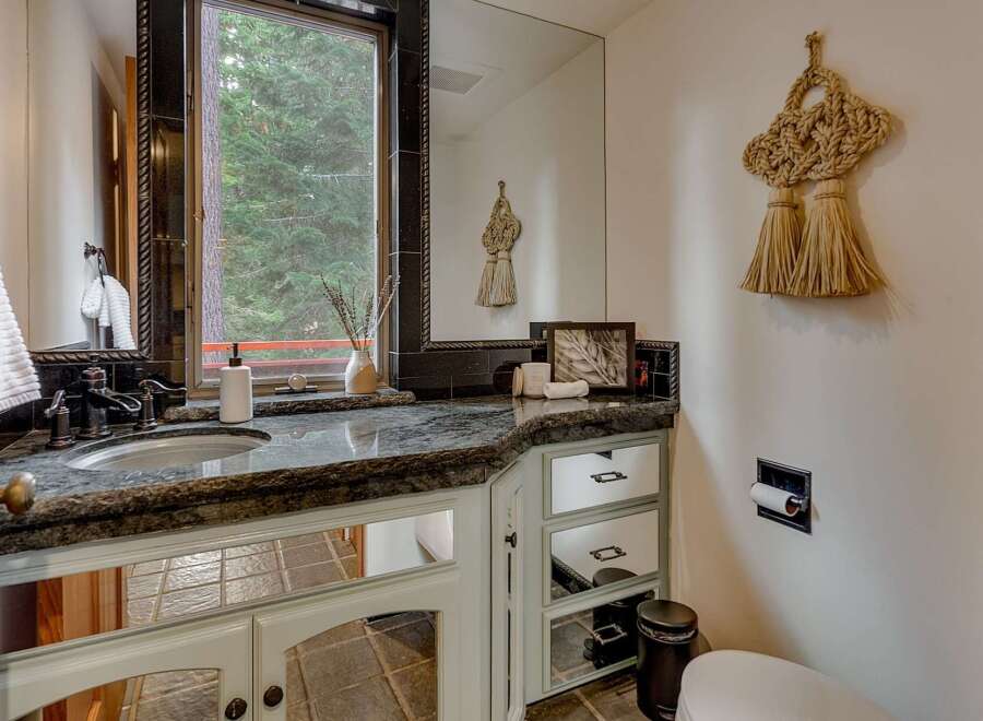 This charming bathroom features a granite countertop with an undermount sink and dark bronze fixtures, complemented by a large mirror reflecting the serene forest view outside the window.