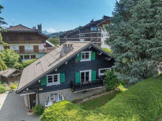 Charming chalet-style home featuring dark wood siding and vibrant green shutters, set against a backdrop of lush greenery and distant mountains.