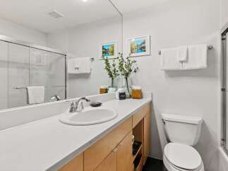 This modern bathroom features a sleek vanity with a white countertop and light wood cabinetry, complemented by a large mirror reflecting the tiled shower and a decorative wall art.