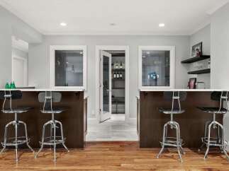 This modern basement bar area features two dark wood bar counters with white countertops, each equipped with four sleek chrome and black stools.